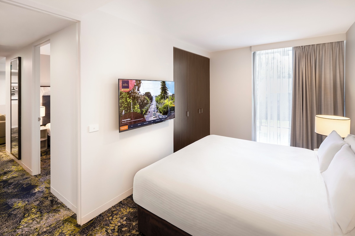 A hotel room with a bed, a TV mounted on the wall, a window with curtains, and a lamp.