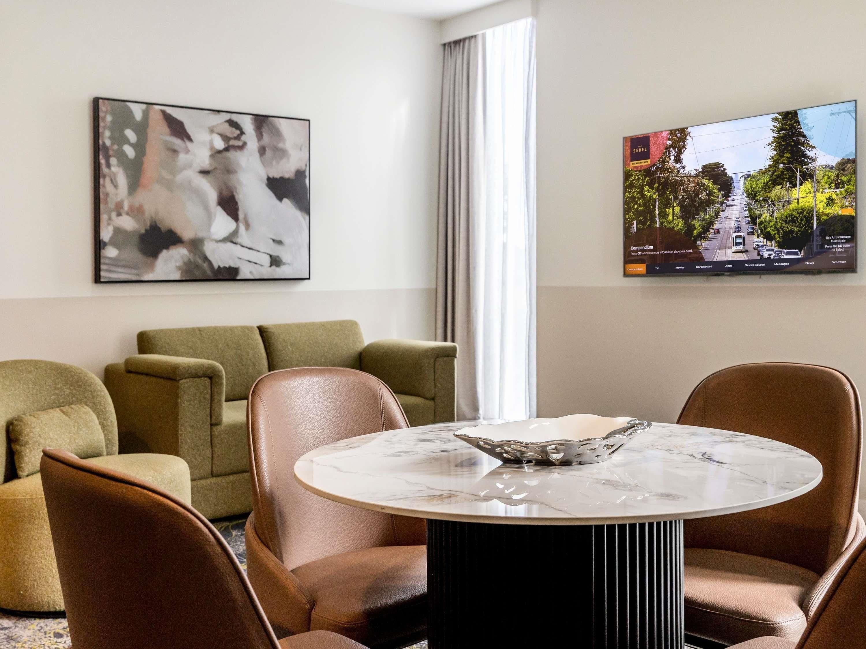 A modern hotel room with a round marble table, leather armchairs, a green sofa, a large abstract painting, and a television displaying a cityscape.