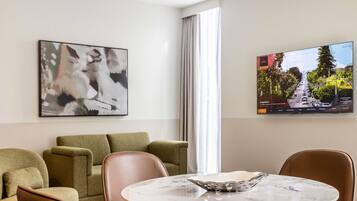 A modern hotel room with a round marble table, leather armchairs, a green sofa, a large abstract painting, and a television displaying a cityscape.