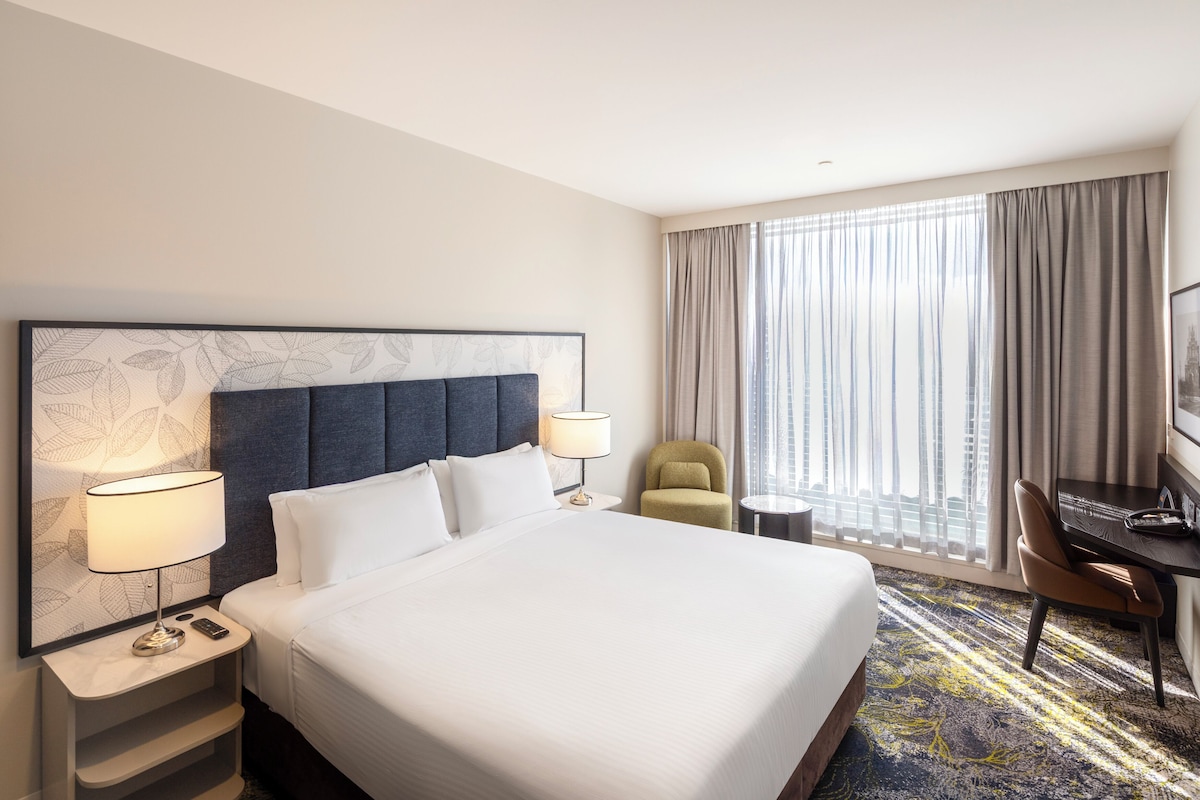 A hotel room with a large bed, two bedside tables with lamps, a desk, a chair, and a view of the city through large windows.