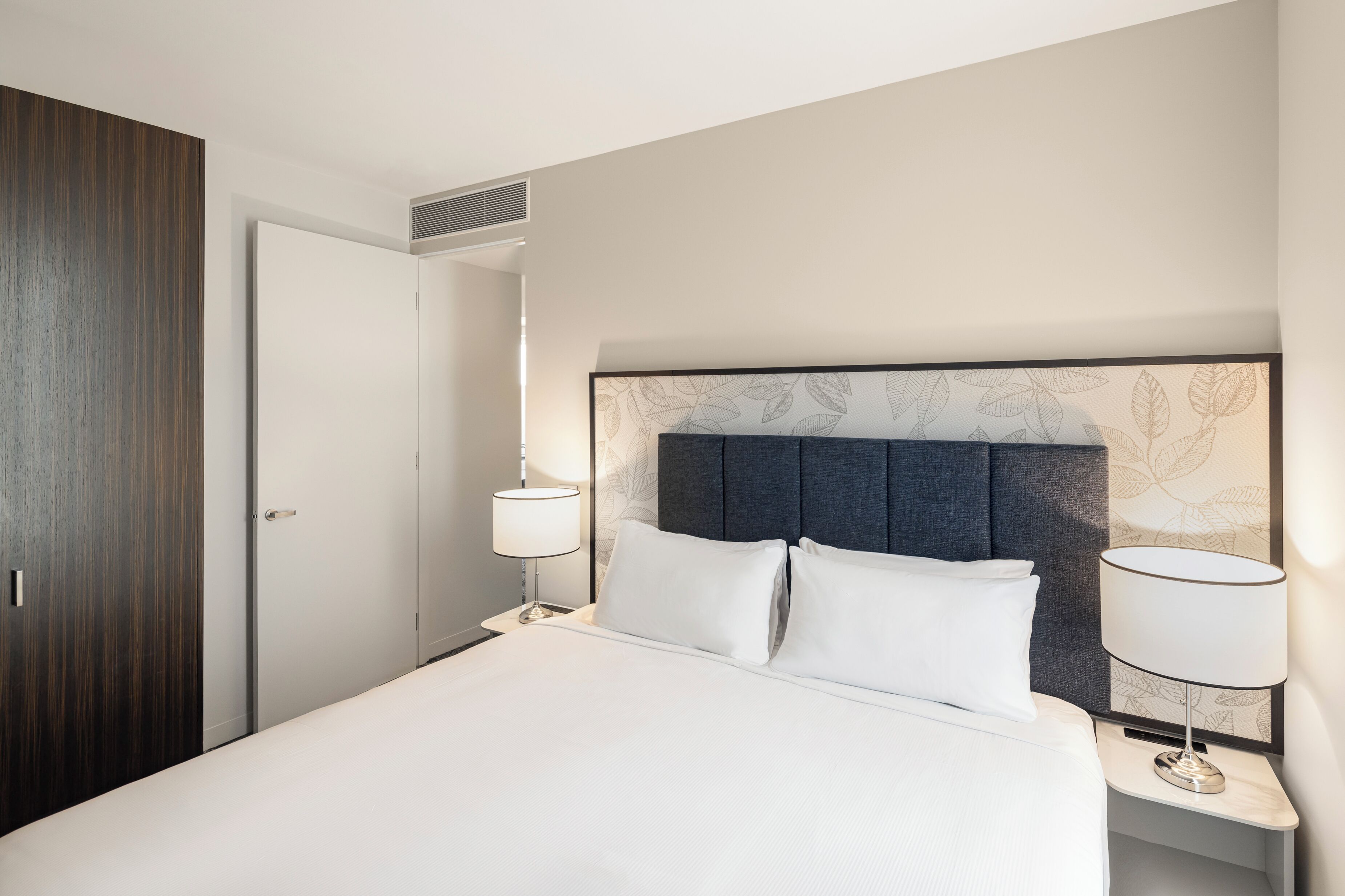 Una habitación de hotel con cama, dos lámparas de noche, cabecera con diseño de hojas, armario de madera y una puerta.