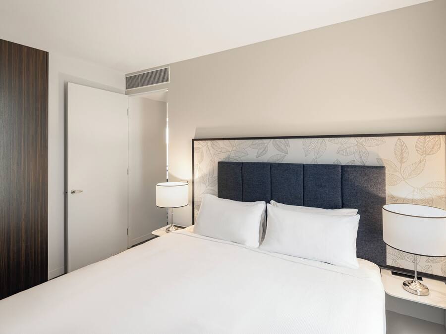 Una habitación de hotel con cama, dos lámparas de noche, cabecera con diseño de hojas, armario de madera y una puerta.