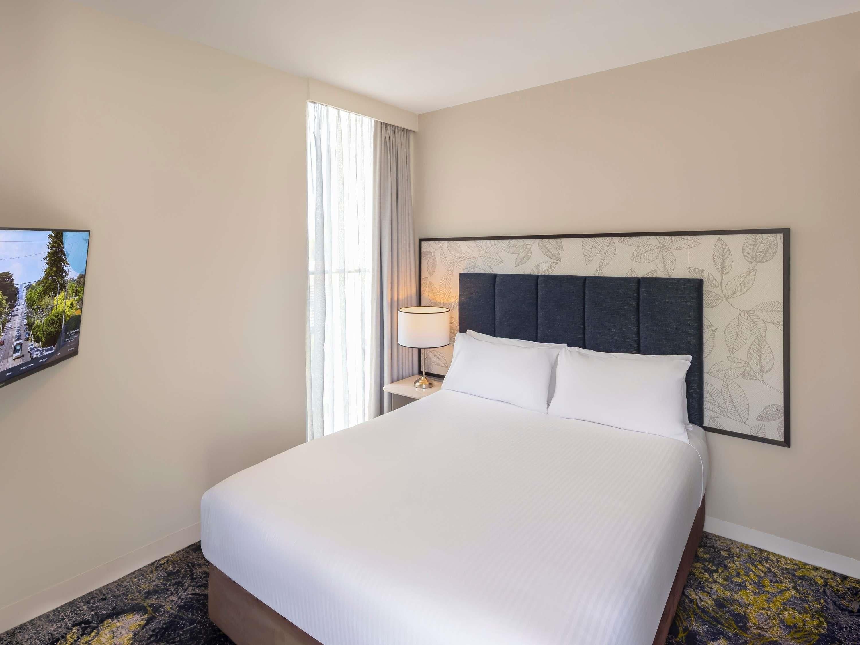 A hotel room with a large bed, a nightstand with a lamp, a wall-mounted TV, and a window with curtains.
