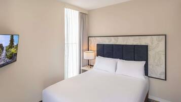 A hotel room with a large bed, a nightstand with a lamp, a wall-mounted TV, and a window with curtains.