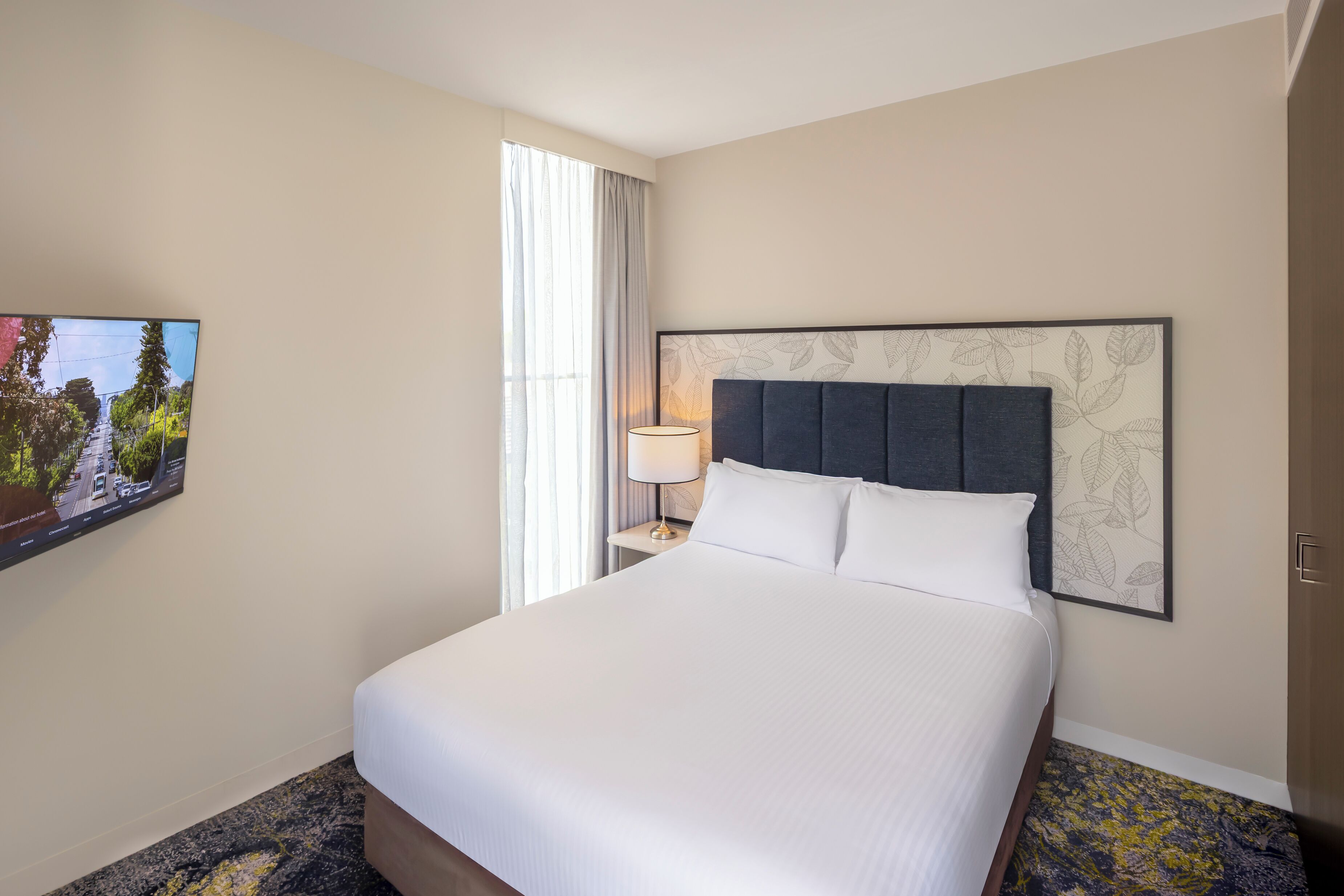 A hotel room with a large bed, a flat-screen TV mounted on the wall, a bedside table with a lamp, and a window with curtains.