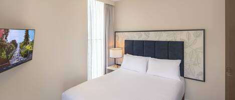 A hotel room with a large bed, a flat-screen TV mounted on the wall, a bedside table with a lamp, and a window with curtains.