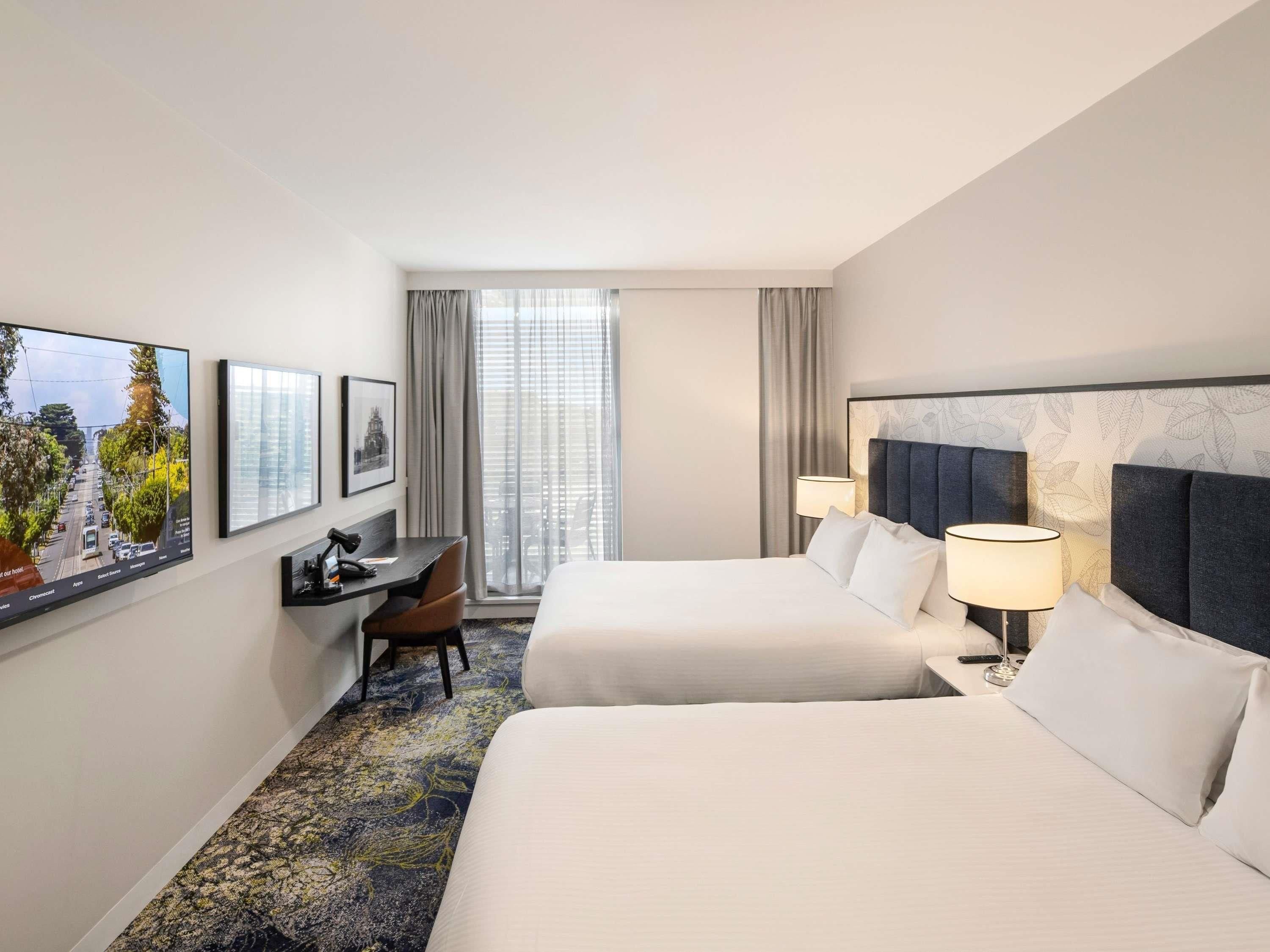 A hotel room with two beds, a desk, a chair, a TV, and a window with curtains.