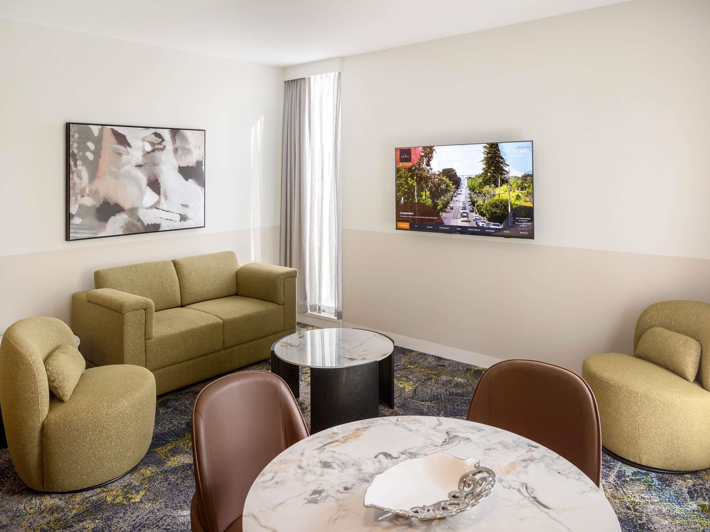 A hotel room with a sofa, armchairs, a small table, a TV, and a window with curtains.