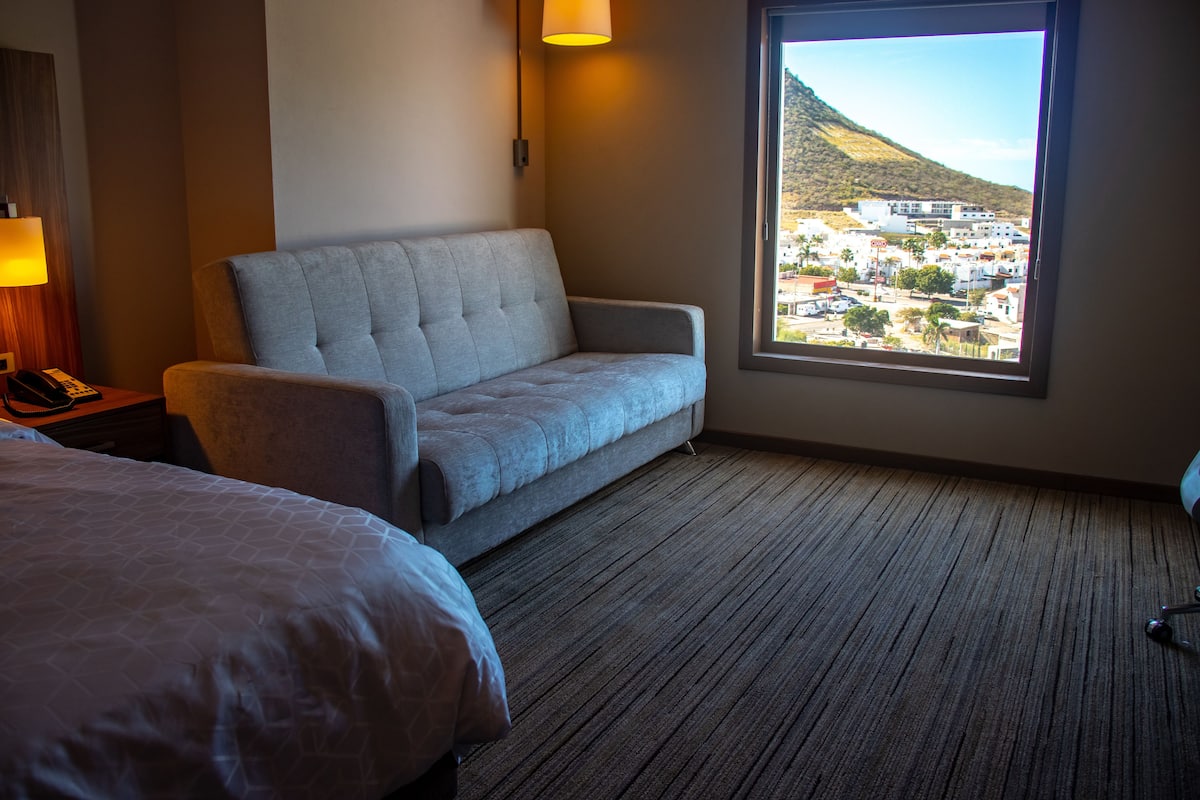 Habitación de hotel con sofá gris, cama con colcha estampada, suelo alfombrado y ventana que ofrece vistas a un pueblo y colinas.