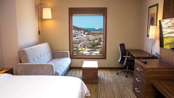 Habitación de hotel con cama, sofá, escritorio con silla, televisión y ventana grande que ofrece vistas a un pueblo costero.