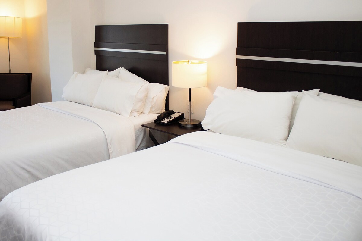 Quarto de hotel com duas camas, uma cadeira, uma mesa de cabeceira com abajur e um telefone.
