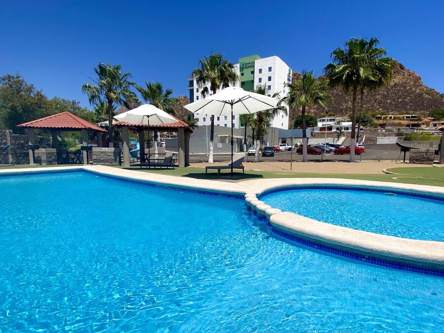 Una piscina con lettini e ombrelloni, con un edificio di un hotel sullo sfondo e un paesaggio collinare.