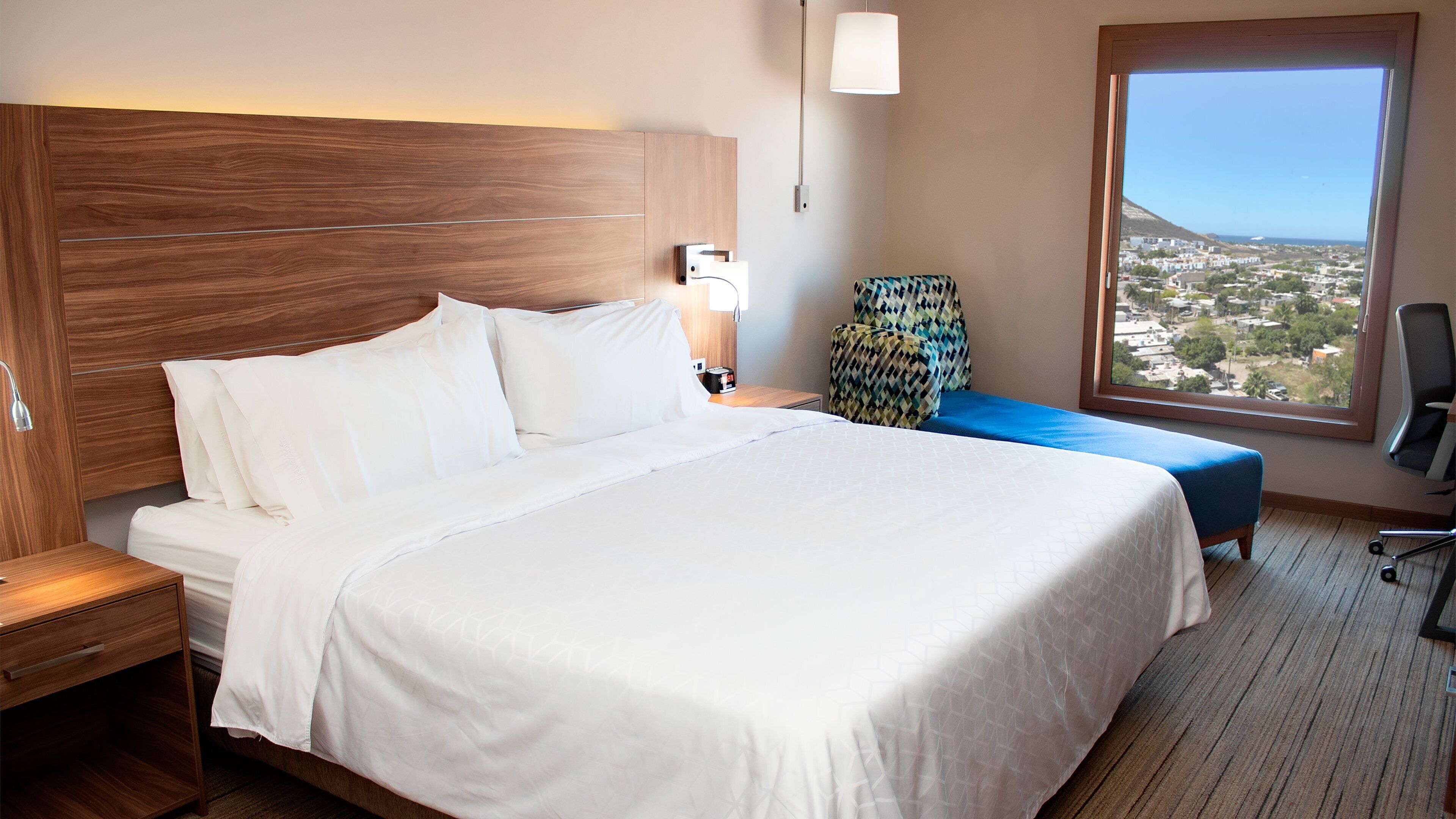 Una habitación de hotel con una cama grande, cabecera de madera, mesita de noche, lámpara, silla y vista a un paisaje urbano a través de una ventana.