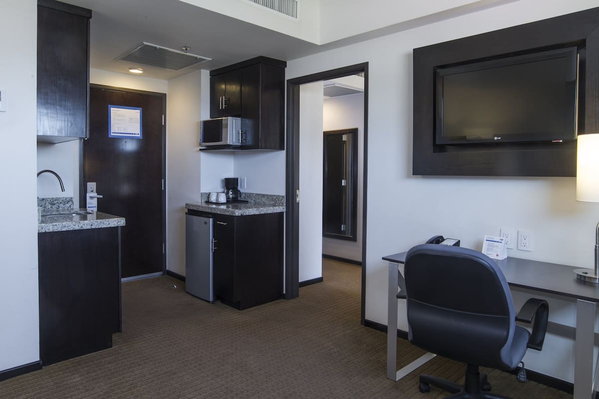 Habitación de hotel compacta con kitchenette, un escritorio con silla, televisor de pantalla plana y una lámpara.