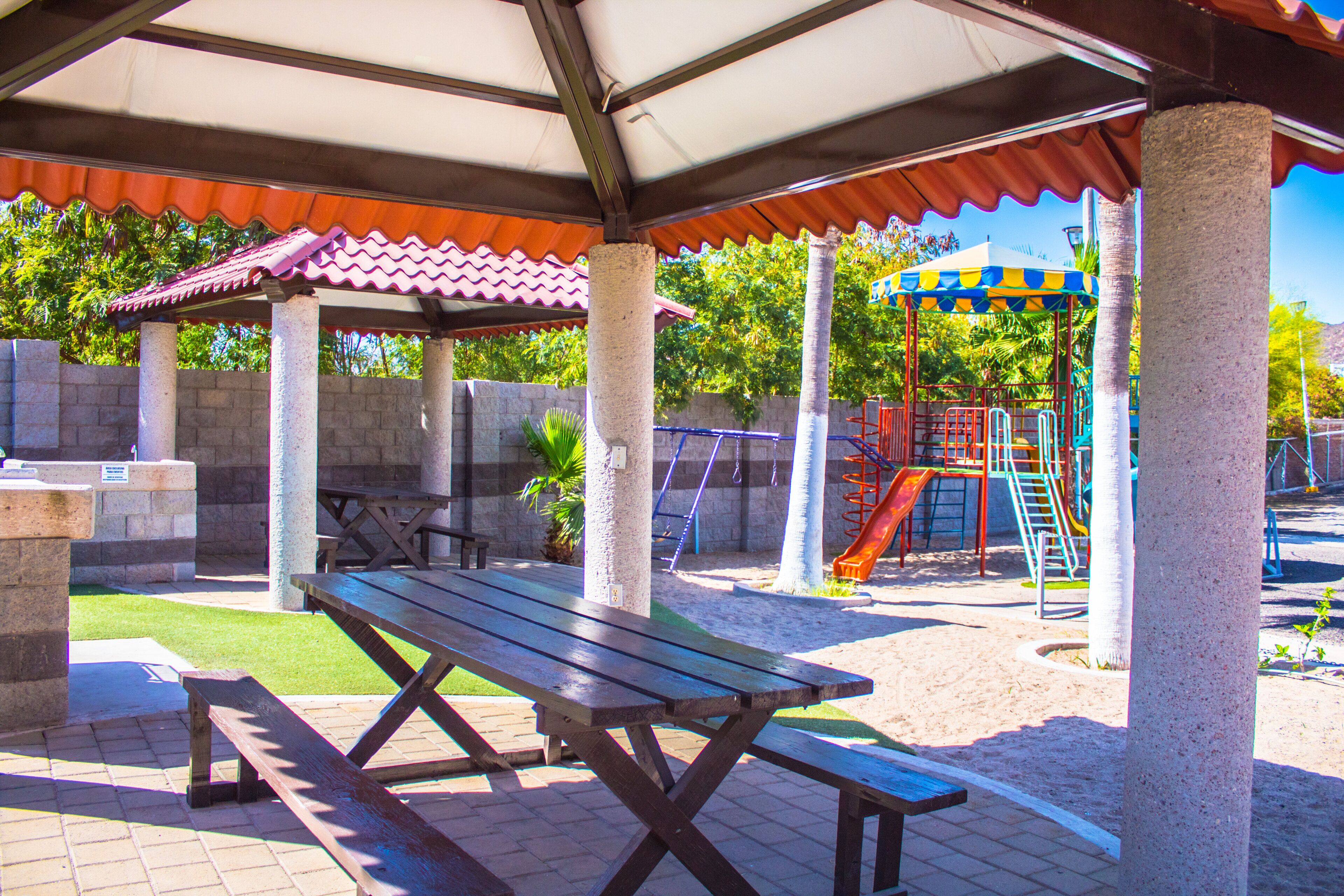 A covered picnic area with benches, a playground with slides, and palm trees.