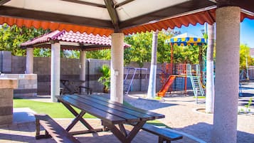 A covered picnic area with benches, a playground with slides, and palm trees.