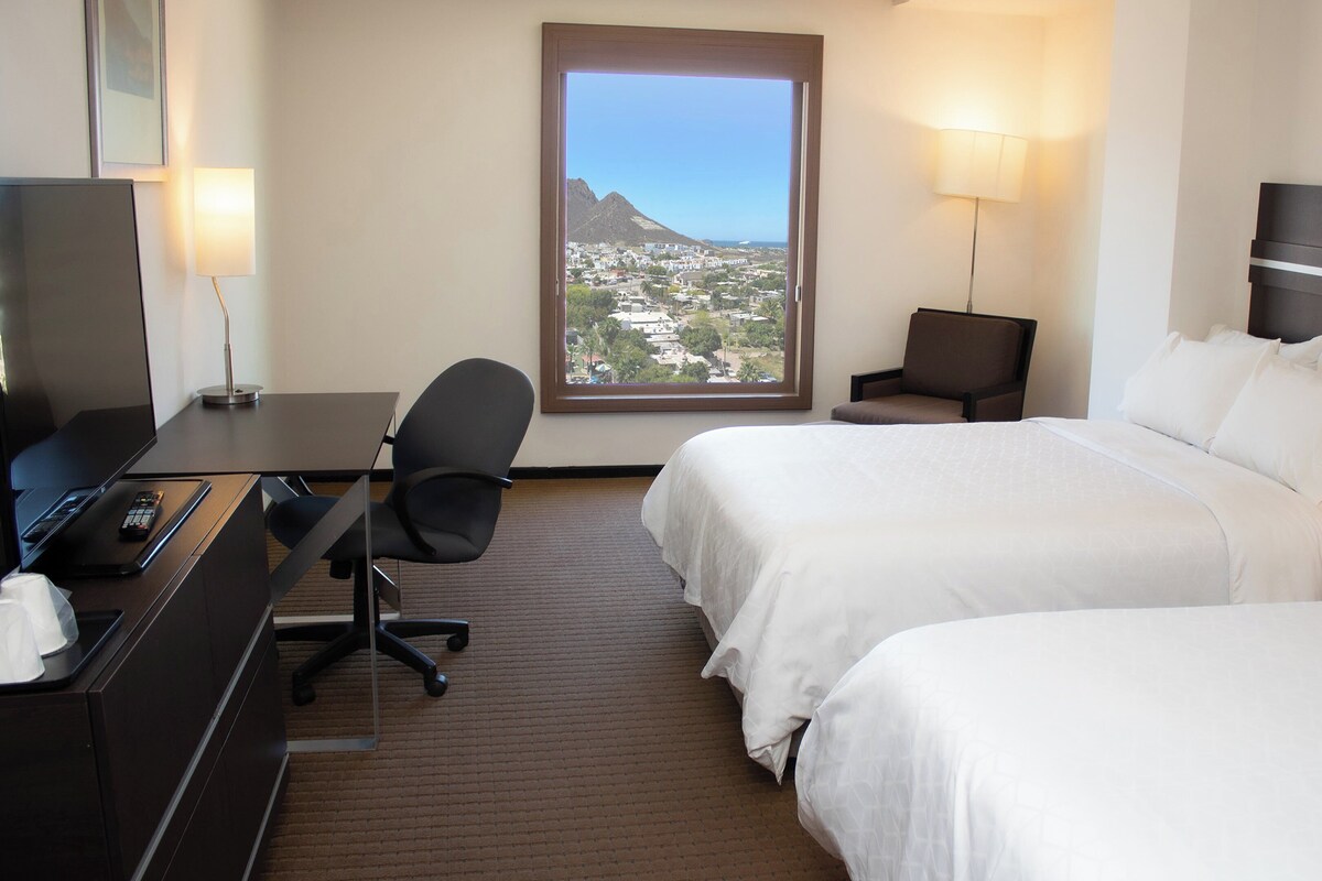 Quarto de hotel com duas camas, uma escrivaninha com computador e comando remoto, uma cadeira, um abajur e vista para a cidade através de uma grande janela.