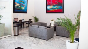 Un salon moderne avec un canapé gris, une table basse et une plante en pot. Deux tableaux abstraits ornent le mur, ainsi qu’un bureau d’ordinateur et une poubelle.