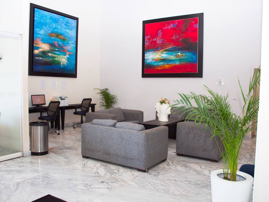 Een moderne woonkamer met een grijze bank, een salontafel en een potplant. Twee abstracte schilderijen hangen aan de muur, een bureau en een prullenbak.