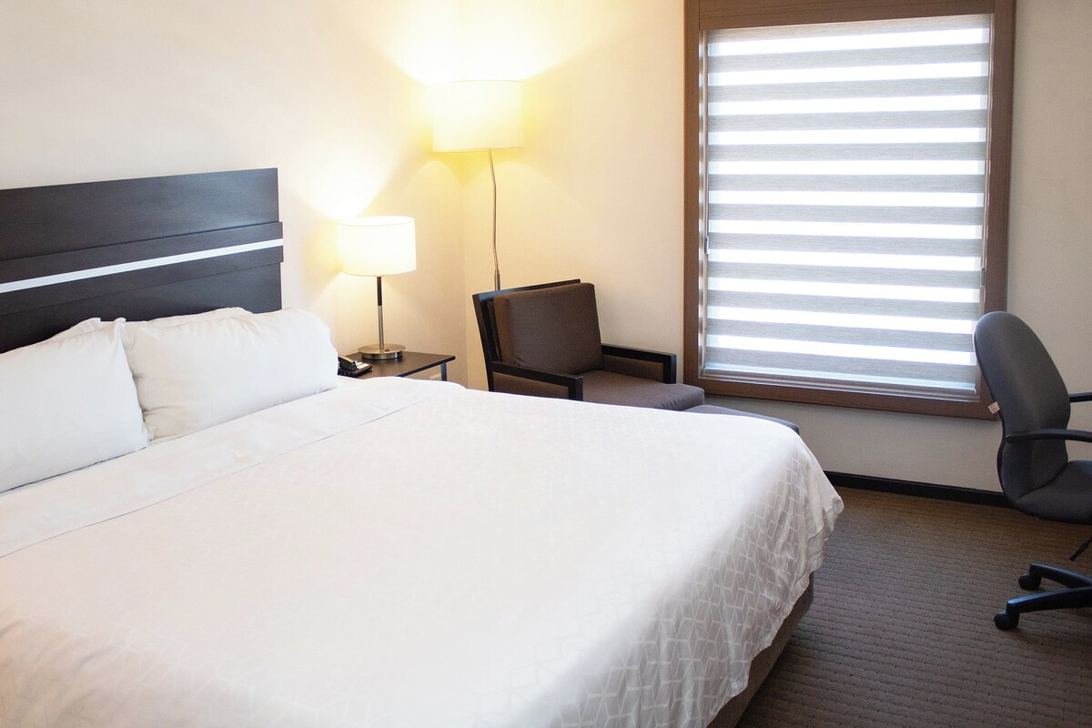 Quarto de hotel com uma cama grande, uma escrivaninha, uma cadeira, um abajur e uma janela com persianas.