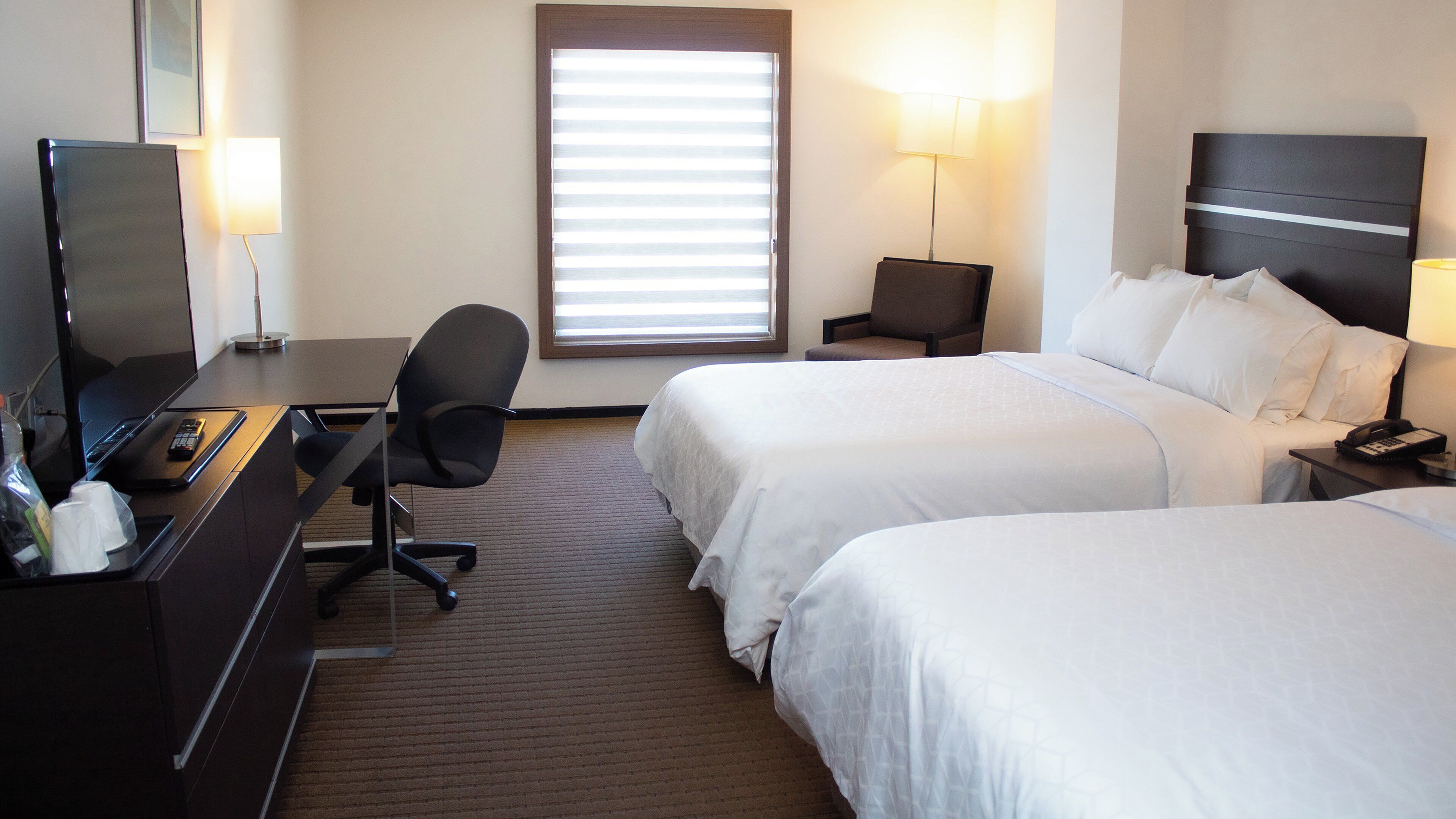 A hotel room with two beds, a desk with a computer, a chair, a TV, and a window with blinds.