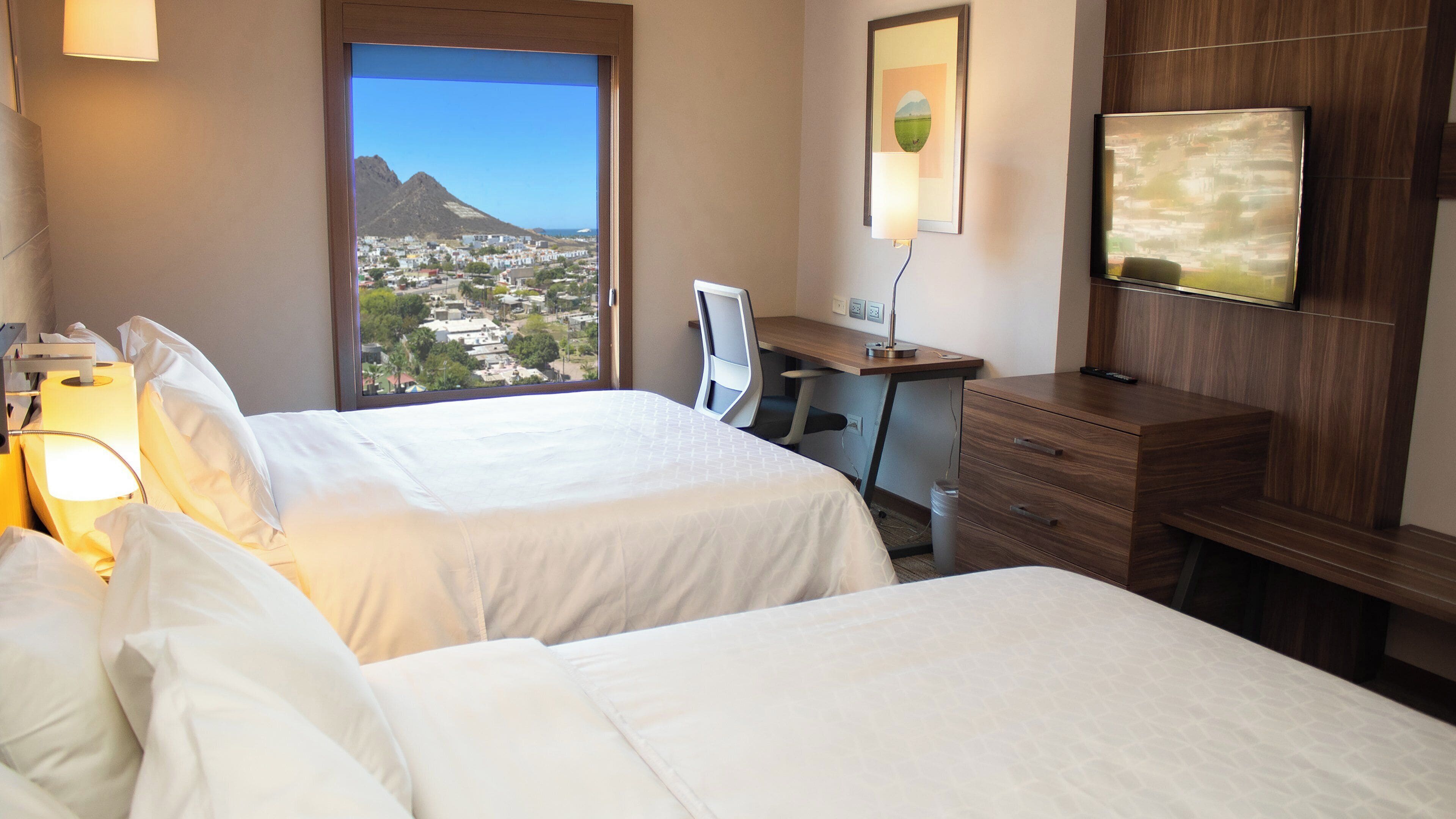 A hotel room with two beds, a desk, a chair, a TV, and a window with a cityscape view.