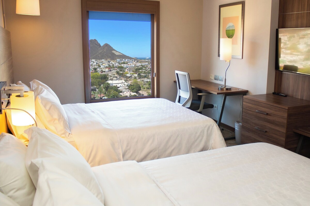 Quarto de hotel com duas camas, uma escrivaninha, uma cadeira, uma TV e uma janela com vista para a cidade.