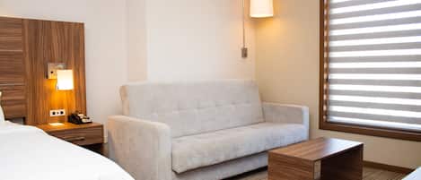 A hotel room with a bed, a sofa, a nightstand with a lamp, a wooden headboard, and a window with blinds.
