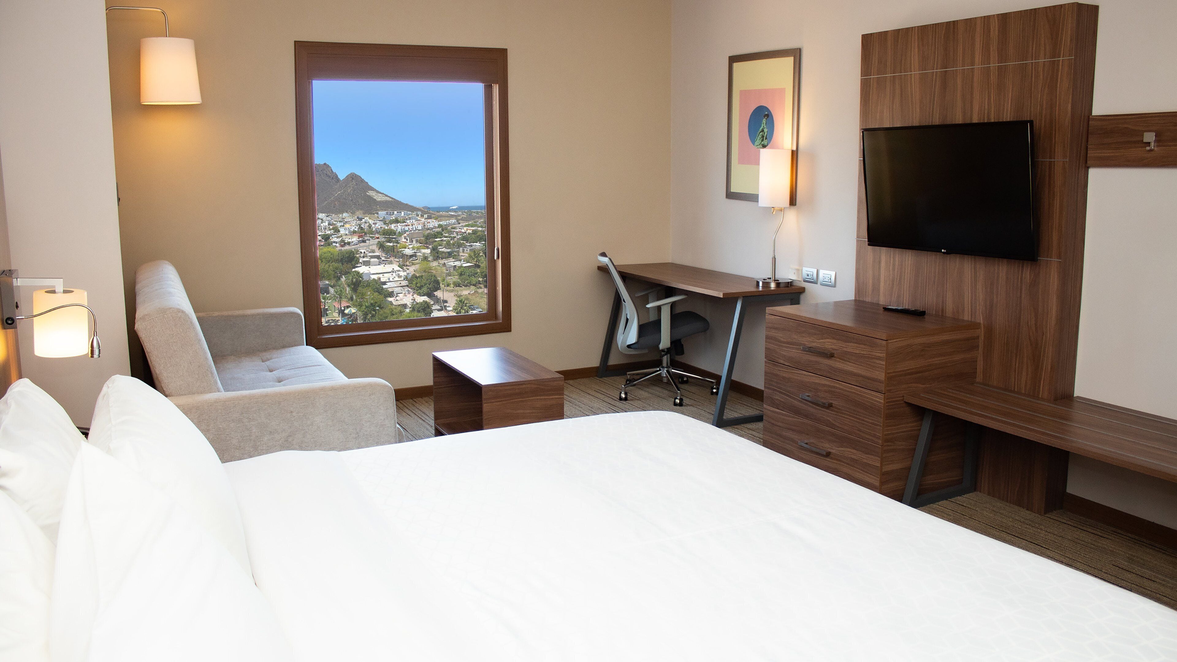 A hotel room with a bed, a desk, a chair, a TV, and a view of a cityscape through a large window.