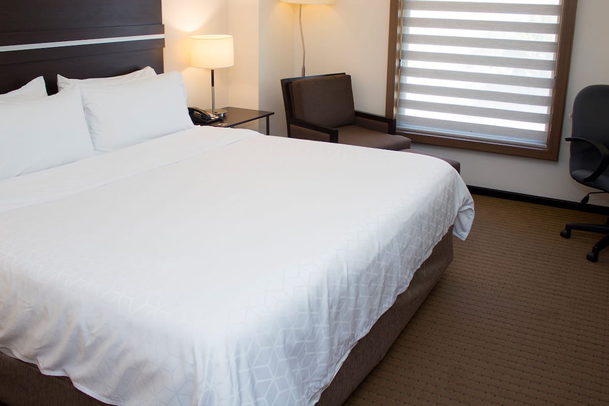 Habitación de hotel con una cama grande, un escritorio con lámpara, una silla y una ventana con persianas.