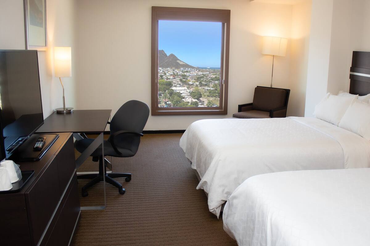 Habitación de hotel con dos camas, un escritorio con computadora y control remoto, una silla, una lámpara y vista panorámica de la ciudad a través de un ventanal grande.