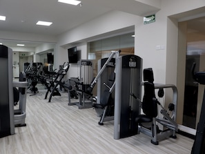 Uma academia com diversos equipamentos de exercício, uma televisão de tela plana e uma placa indicando uma rota de evacuação.