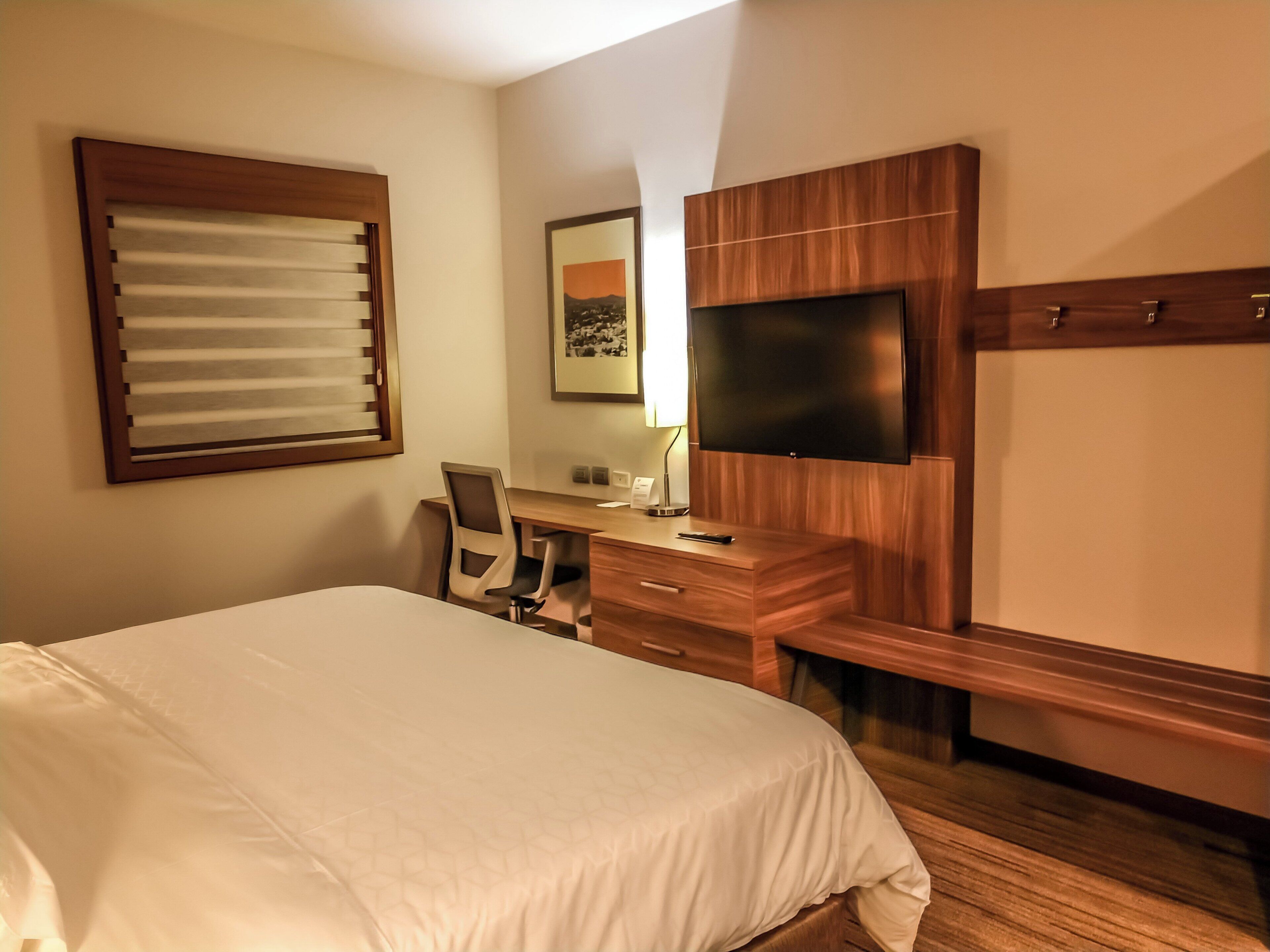 Habitación de hotel con cama, escritorio, silla, televisor y ventana con persianas.