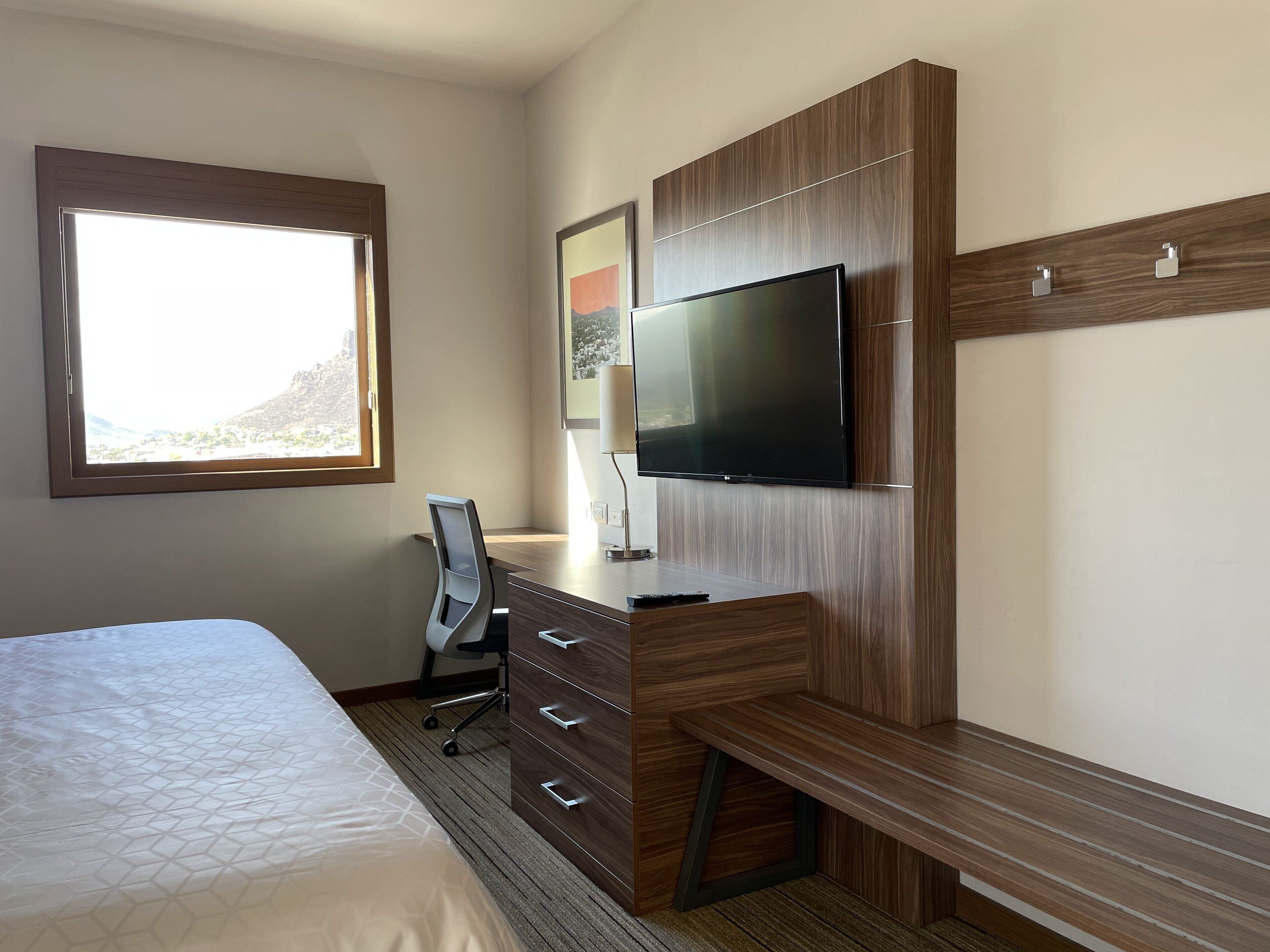 A hotel room with a bed, a desk with drawers, a chair, a TV mounted on a wooden wall, and a window with a view of a mountain.