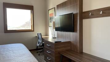 A hotel room with a bed, a desk with drawers, a chair, a TV mounted on a wooden wall, and a window with a view of a mountain.