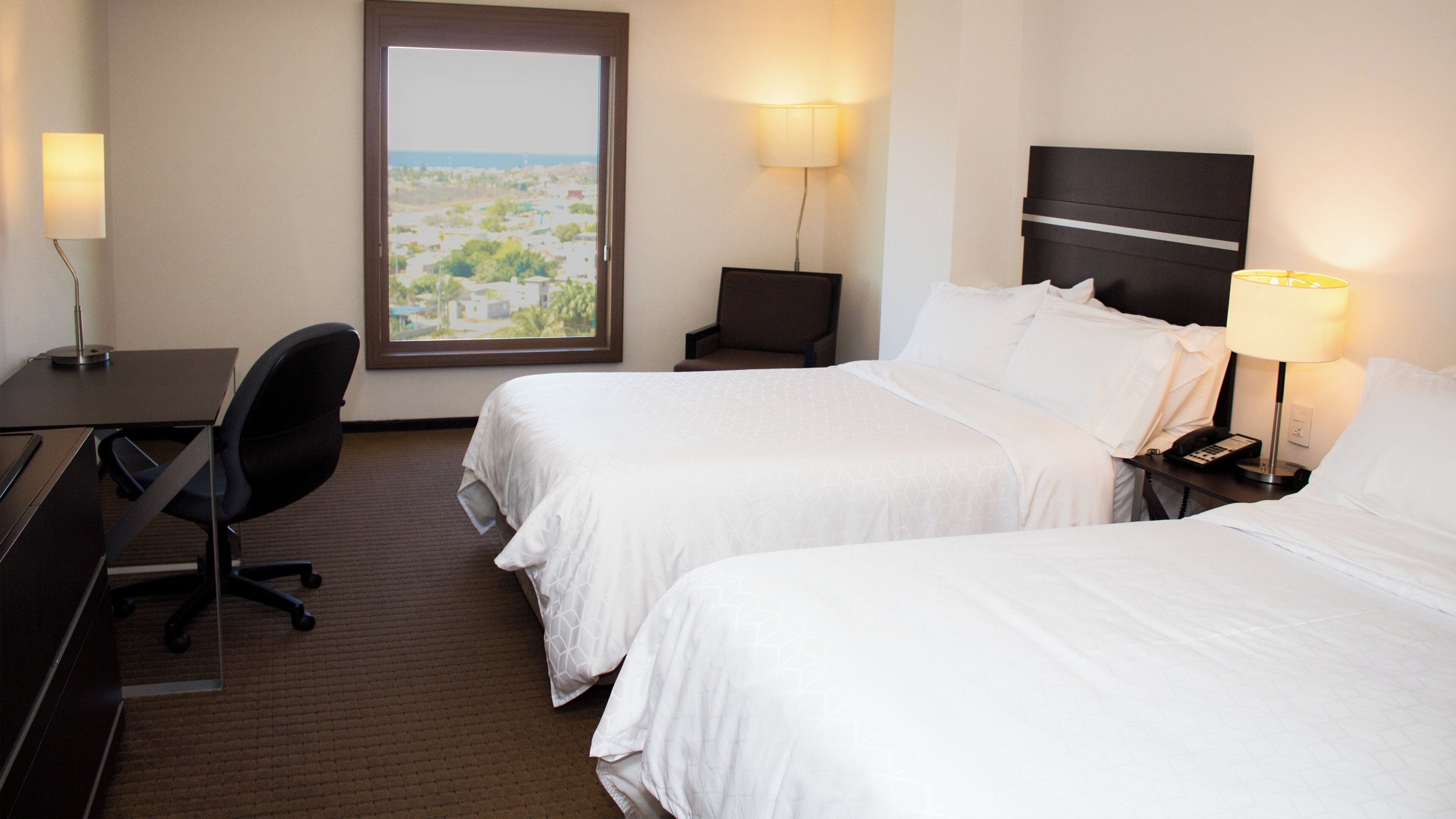 A hotel room with two beds, a desk, a chair, and a view of the cityscape.