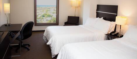 A hotel room with two beds, a desk, a chair, and a view of the cityscape.