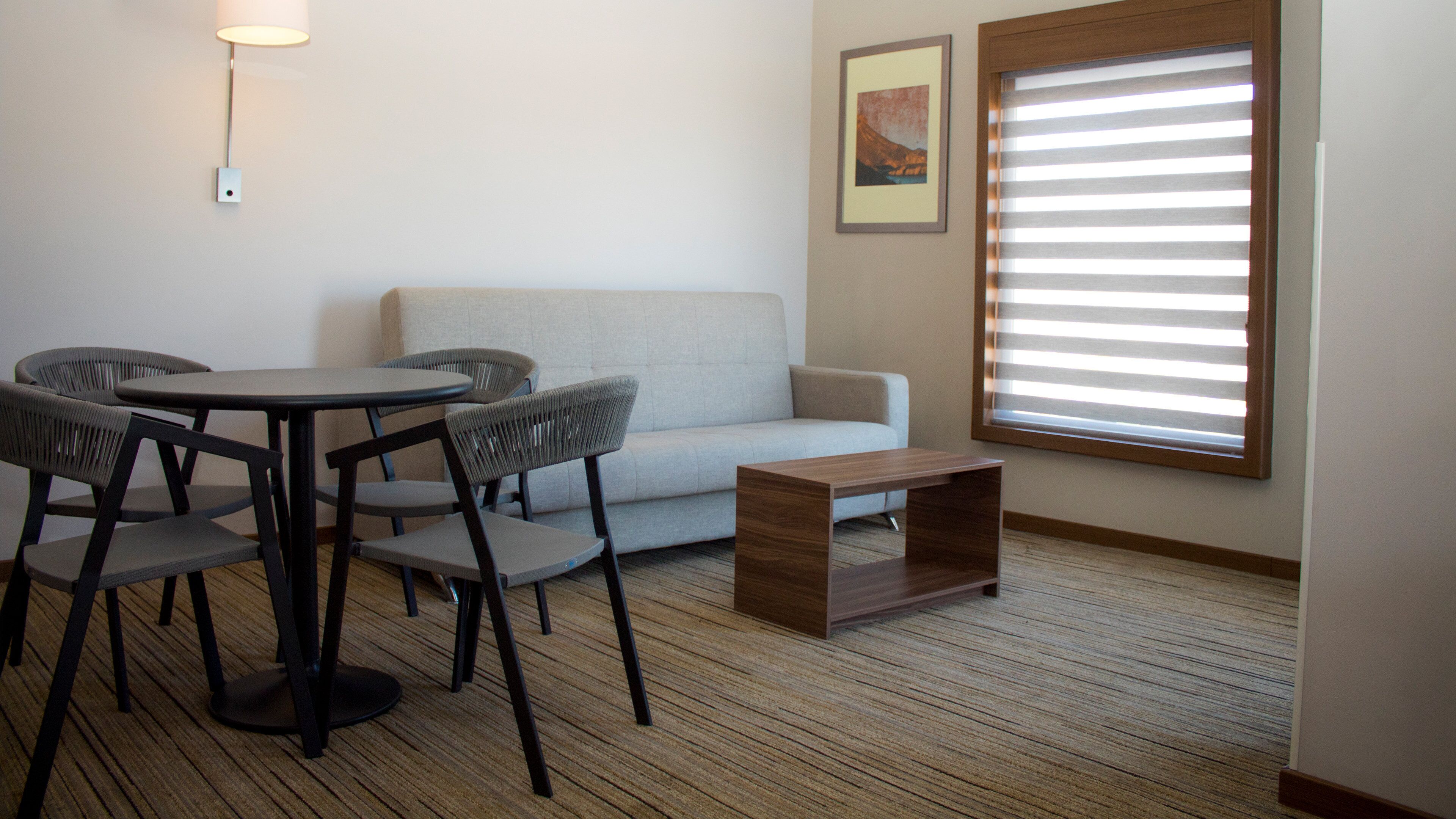Un salon moderne comprenant un canapé, une table ronde avec des chaises, une petite table basse en bois, une lampe et une fenêtre avec des stores.