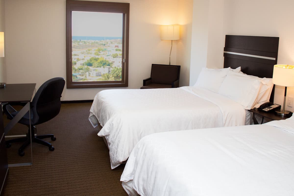 Habitación de hotel con dos camas, un escritorio, una silla y una ventana con vista a la ciudad.