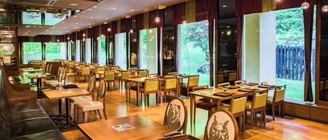 A restaurant with multiple dining tables and booths, large windows with a view of greenery, and hanging pendant lights.
