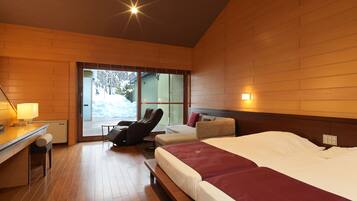 A hotel room with a wooden floor, a bed with a red blanket, a massage chair, a desk with a lamp, and a view of a snowy landscape through the window.