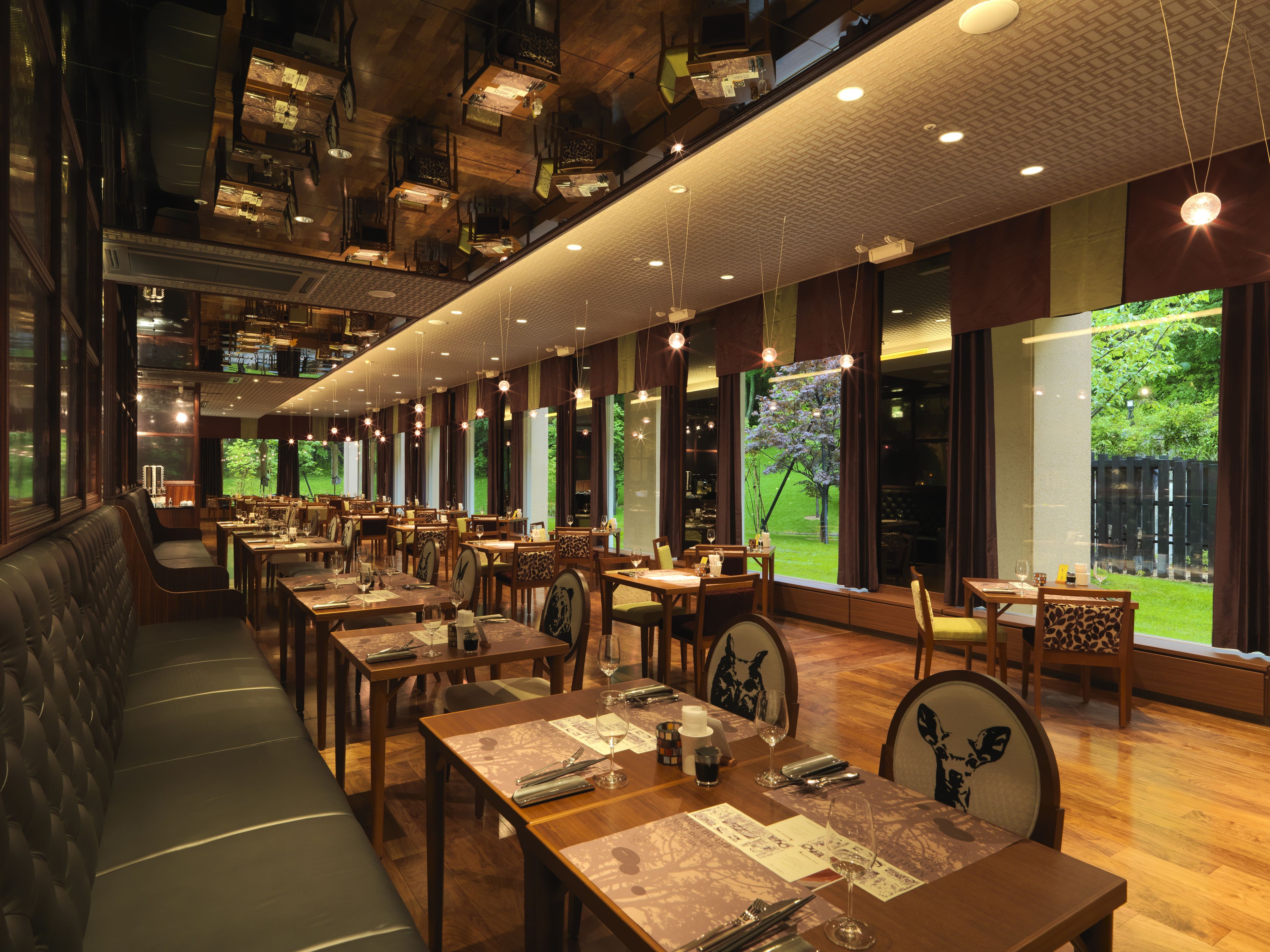 Un restaurante con varias mesas y cabinas, amplios ventanales con vista a la vegetación y lámparas colgantes.