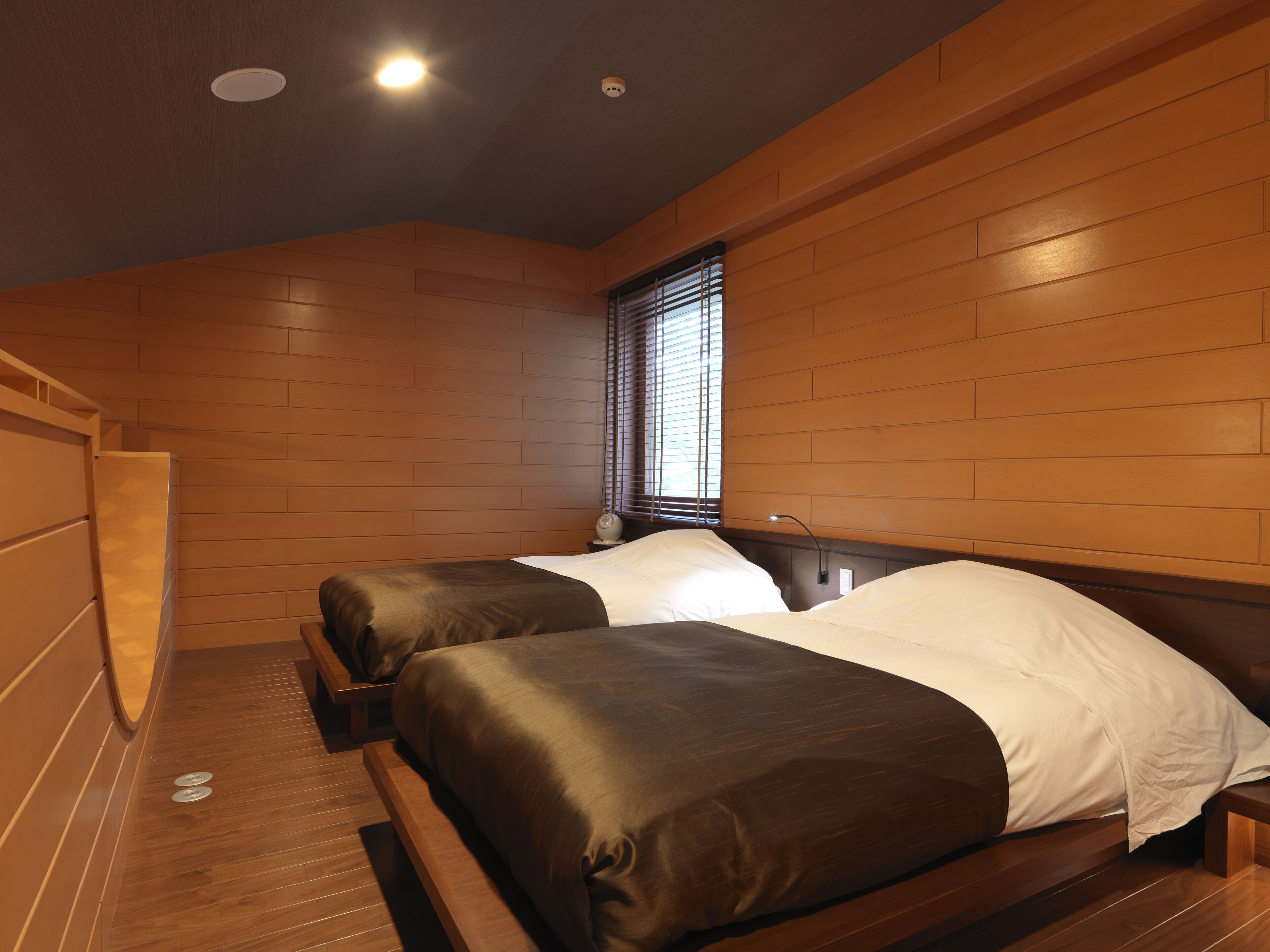 A room with two beds, wooden walls, a window with blinds, and a ceiling light.