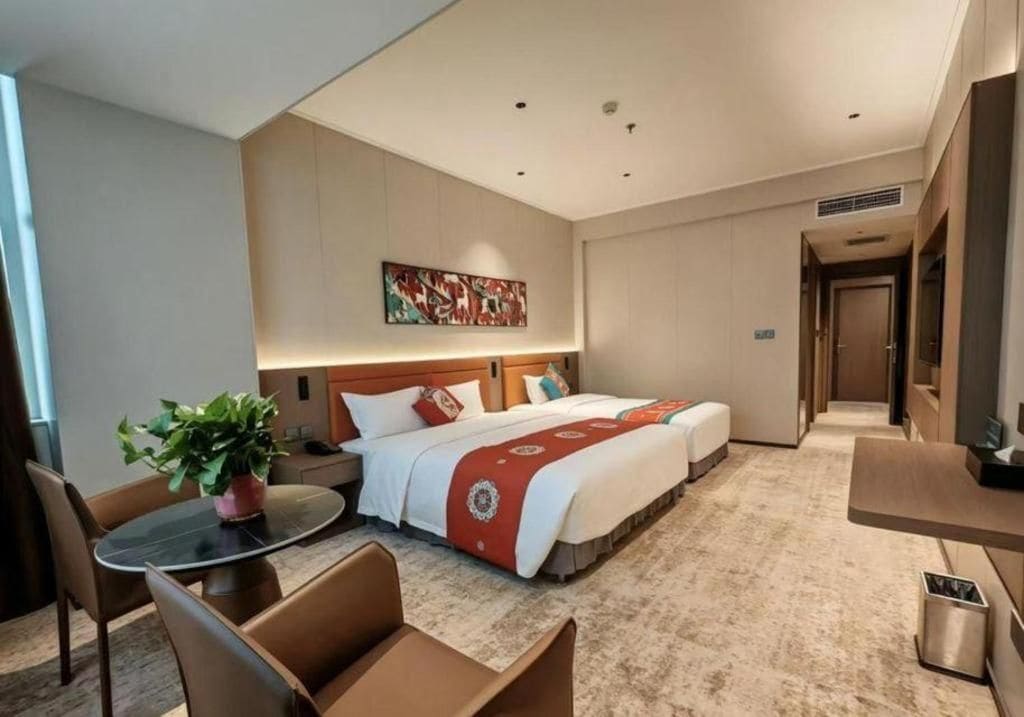 A hotel room with a bed, a desk, a chair, a table, and a potted plant.
