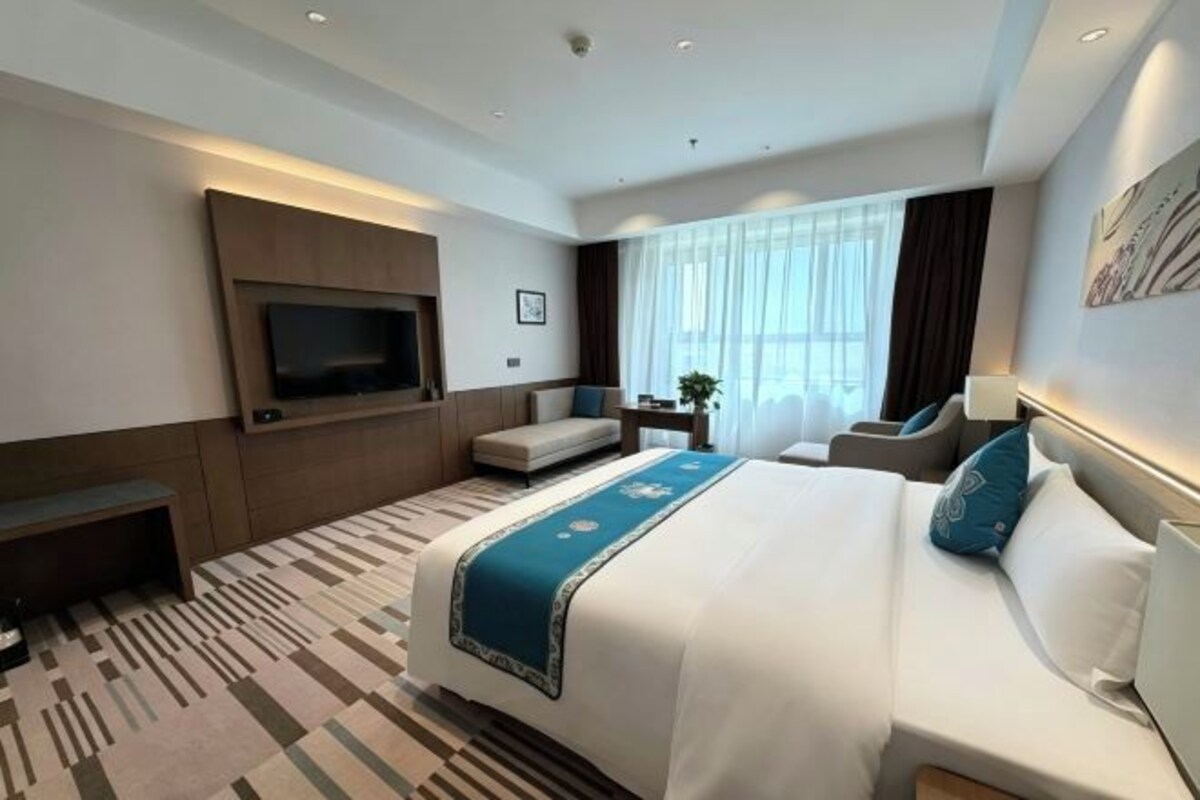 Habitación de hotel con una cama grande, un televisor de pantalla plana montado en la pared, una zona de estar con un banco y una ventana con cortinas.