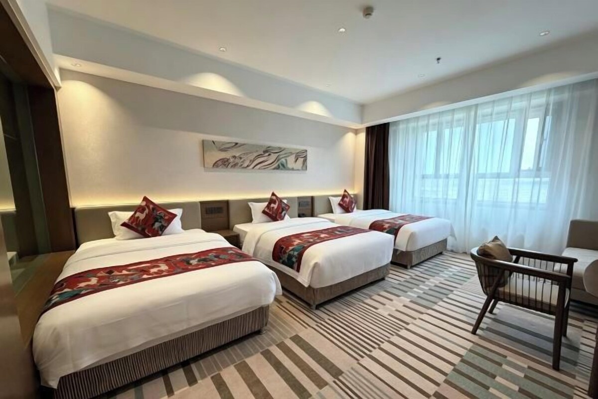 A hotel room with three beds, a large window, a wall art piece, and a patterned rug.