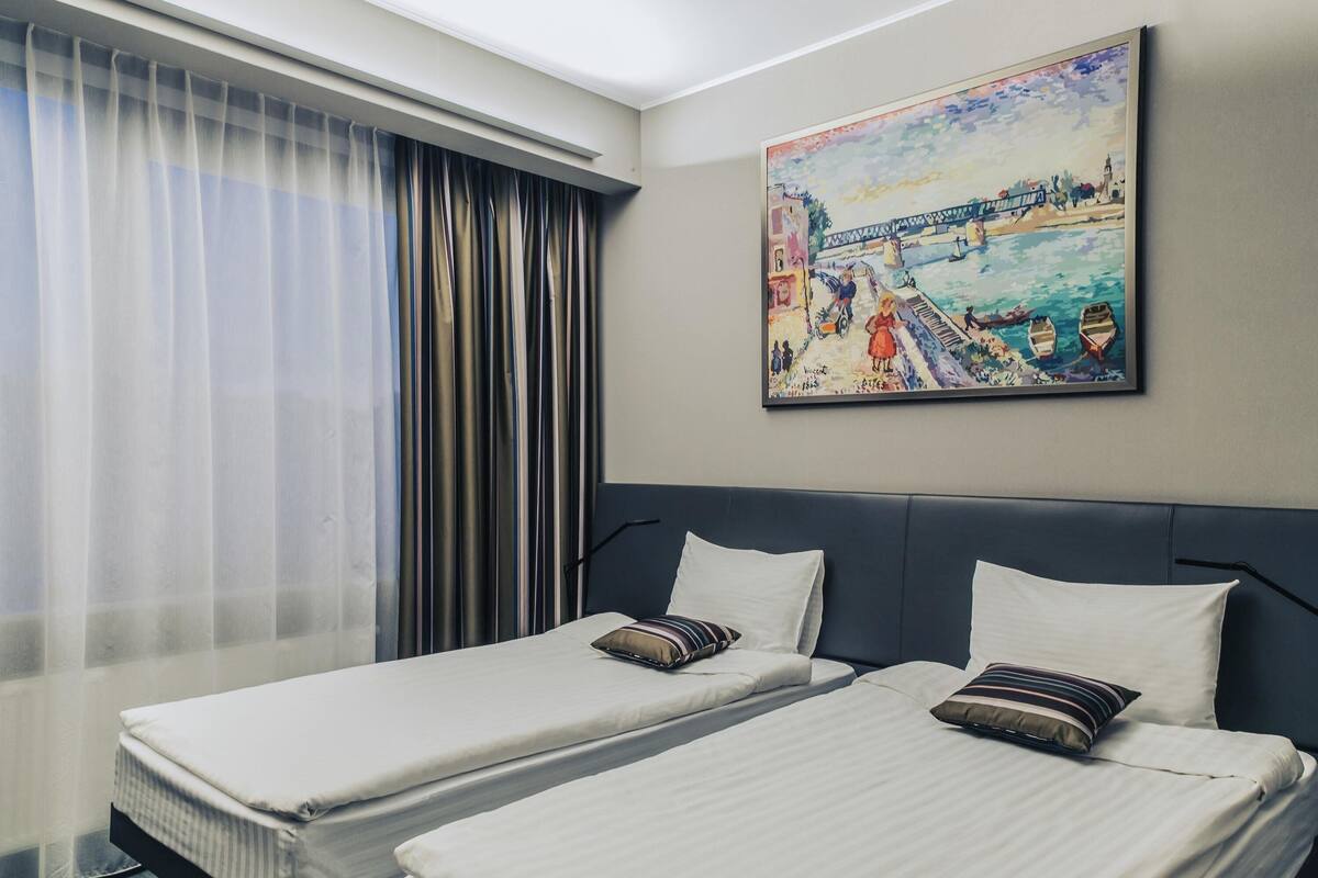 Ett hotellrum med två separata sängar, ett stort fönster med tunna, genomskinliga gardiner och en tavla som föreställer ett kustlandskap på väggen.
