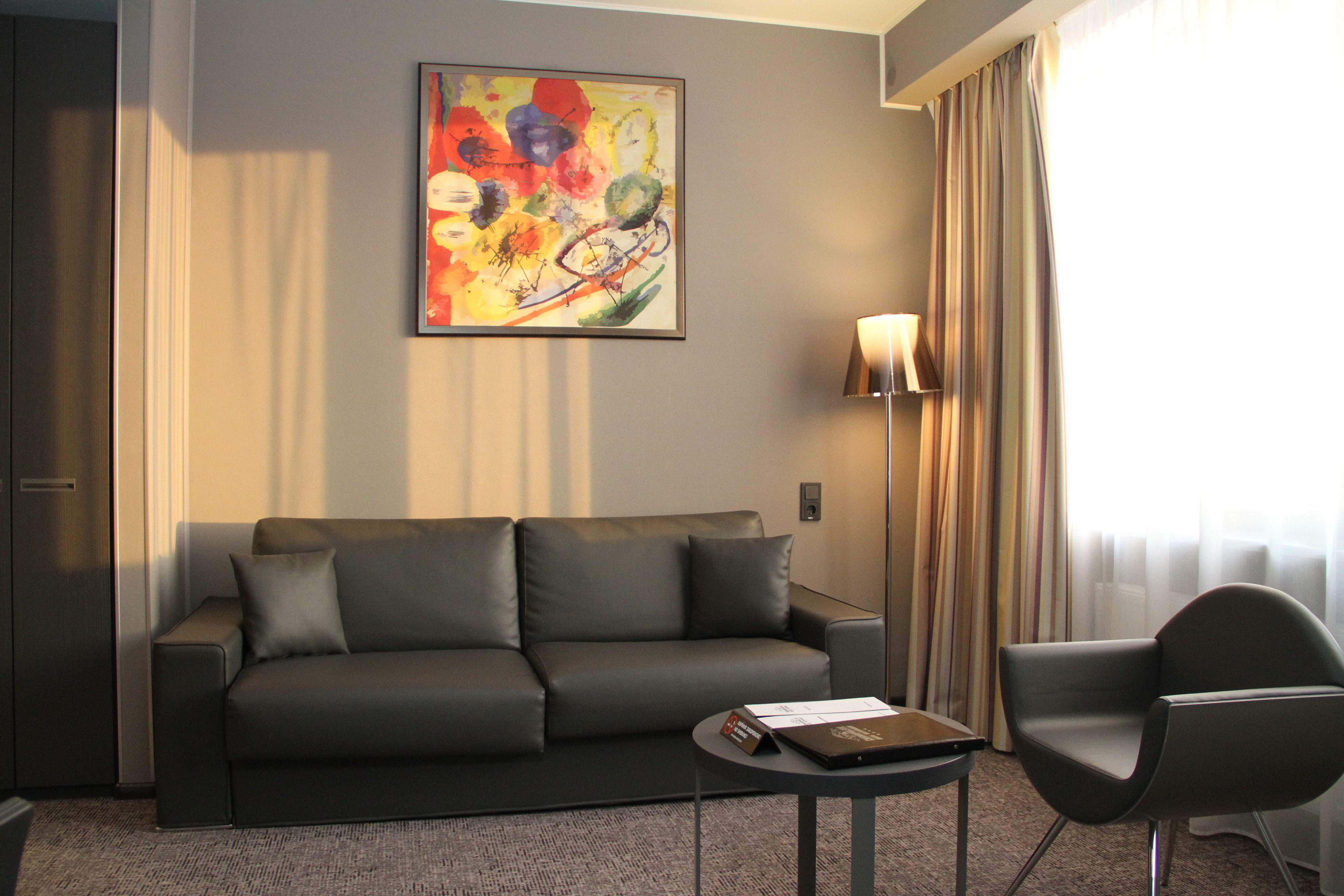Moderni hotellihuone, jossa on harmaa sohva, pieni pyöreä pöytä, yksi nojatuoli, värikäs abstrakti maalaus seinällä ja suuri ikkuna, jossa on verhot.