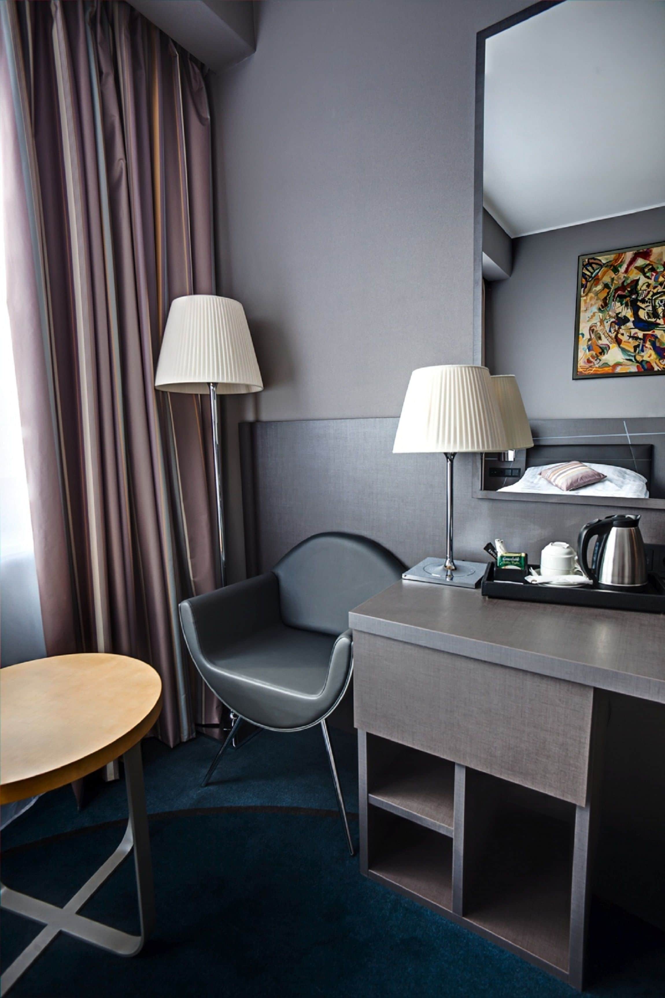 A modern hotel room with a desk, chair, lamp, and a painting on the wall.
