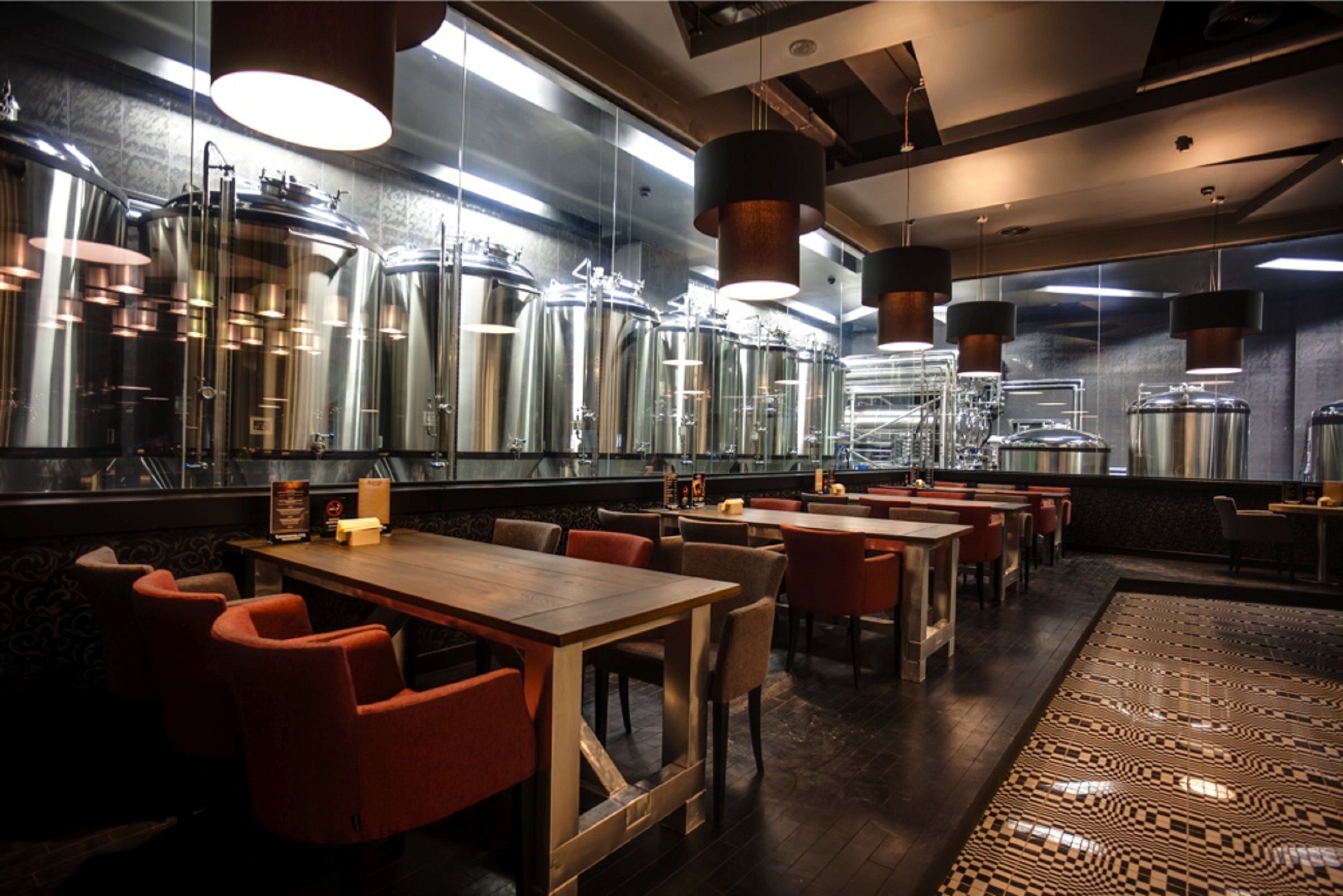 Un restaurante con varias mesas y sillas, tanques de elaboración industrial a gran escala al fondo y un suelo con estampado.